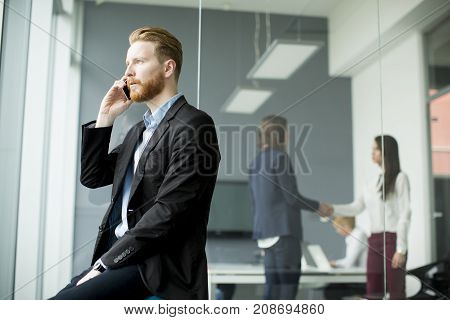 Confident businessman with ginger hair using mobile phone while other business people working in office