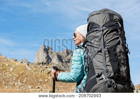 Hiker girl in sunglasses down jacket and hat with a backpack and mountain equipment with tracking sticks in her hands looks at the beautiful view of a high mountain lake in an archipelago in the northern Caucasus. Karachay-Cherkess Republic. 