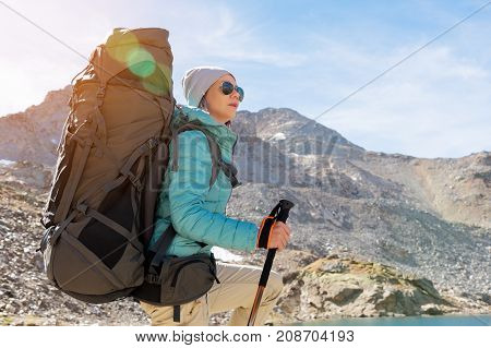 Hiker girl in sunglasses down jacket and hat with a backpack and mountain equipment with tracking sticks in her hands looks at the beautiful view of a high mountain lake in an archipelago in the northern Caucasus. Karachay-Cherkess Republic. 