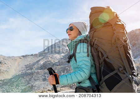Hiker girl in sunglasses down jacket and hat with a backpack and mountain equipment with tracking sticks in her hands looks at the beautiful view of a high mountain lake in an archipelago in the northern Caucasus. Karachay-Cherkess Republic. 