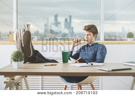Handsome young businessman reading book and drinking coffee in modern office with blurry city view. Education concept