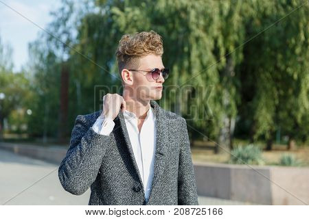 A young guy in the city wearing glasses, against the background of trees in a gray coat. Close-up.