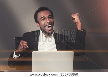 Happy African American businessman is sitting at his laptop in an office and celebrating sealing a good deal. Toned image mock up