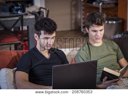 Casual young men relaxing on sofa at home sharing information from textbook and using laptop.