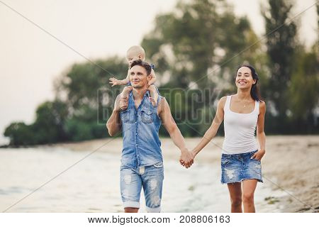 A young family of three walks along a sandy beach. A little baby girl with a blonde one year old from birth sits at the daddy on her shoulders Dressed in blue jeans clothes.