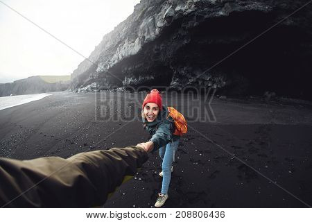 happy smiling woman traveler with small orange backpack walking on Kirkjufjara black sand beach, southern Iceland. a woman holds a man's hand, a man can not be seen, in the frame of his hand