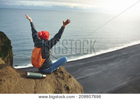 Girl in warm clothing, in red knitted hat and small orange backpack stands on the cliff on background of ocean, sky and Reynisfjara beach in Iceland. Travel to the Iceland