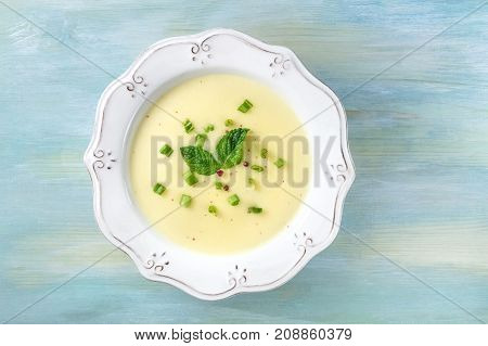 An overhead photo of vichyssoise, a French potato and leeks soup, with chopped green onions, pink peppercorns, and mint leaves for decoration, on a teal background with a place for text