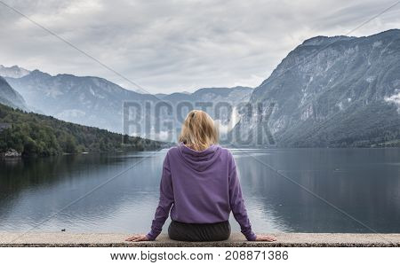 Solitary woman wearing purple hoodie watching tranquil overcast morning scene at lake Bohinj, Alps mountains, Slovenia.
