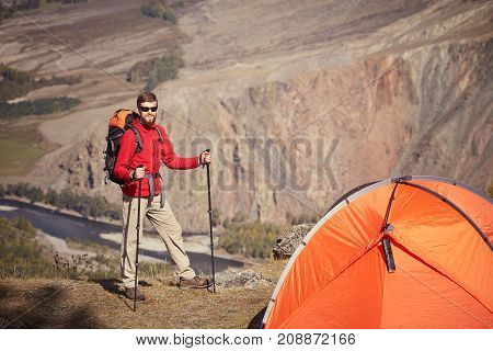 Tall backpacker with bierd and sunglasses stand near orange tent. Sunny spring daybreak in rocky mountains. Hiker with backpack stand on rocky view point above valley.