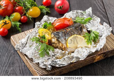 Mackerel baked with tomatoes in foil. Mackerel in foil lies on wooden plate on dark wooden table. Also lie fresh tomatoes and herbs. Healthy and dietary food