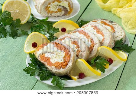 Homemade mackerel roll with carrots and eggs in gelatin. Sliced pieces of rolls are served with lemon cranberries and parsley. Festive snack. Mackerel roll lemon and parsley on green wooden table