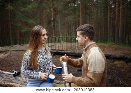 Candid shot of active young couple of travelers having picnic on autumn day in pine wood drinking hot tea or coffee from thermos and having nice conversation enjoying beverage and fresh air