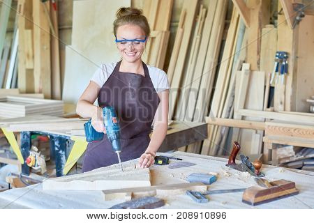 Portrait of cheerful young woman working in carpenters shop drilling holes in piece of wood while making furniture, copy space