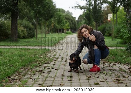Portrait of fashionable young female dog lover in trendy ripped jeans red sneakers and leather jacket sitting on her hunkers on pavement and stroking her four legged friend while walking in park