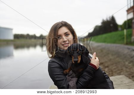 Beautiful blue eyed young European female wearing nose ring and stylish leather jacket posing with dachshund pet dog on her hands enjoying nice walk by the river looking and smiling at camera