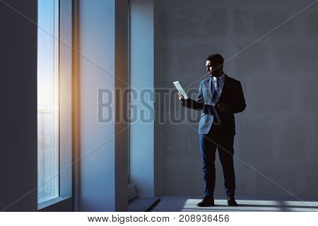 Estate agent stands with tablet computer near big window in blank new apartment on grey wall background. Business concept