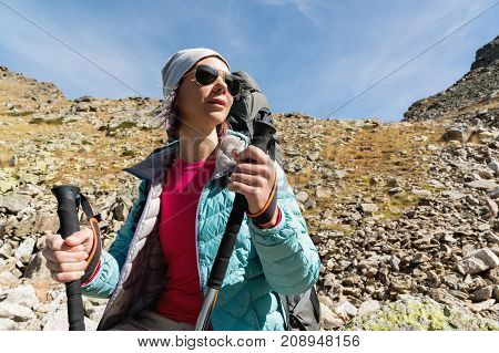 Hiker girl in sunglasses down jacket and hat with a backpack and mountain equipment with tracking sticks in her hands looks at the beautiful view of a high mountain lake in an archipelago in the northern Caucasus. Karachay-Cherkess Republic.