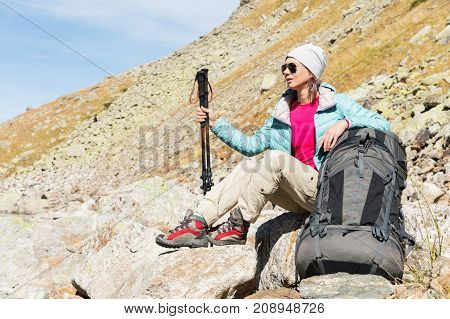 A tourist girl wearing sunglasses down jacket and hat with a backpack and mountain equipment with handles for tracking in her hands is sitting and relaxing looking at the beautiful view of the high mountain lake in the North Caucasus. Karachay-Cherkess 