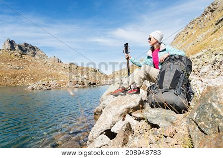 A tourist girl wearing sunglasses down jacket and hat with a backpack and mountain equipment with handles for tracking in her hands is sitting and relaxing looking at the beautiful view of the high mountain lake in the North Caucasus. Karachay-Cherkess 