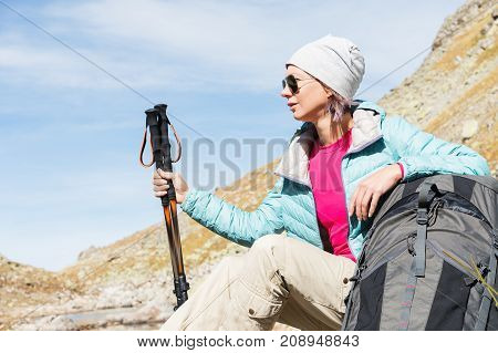 A tourist girl wearing sunglasses down jacket and hat with a backpack and mountain equipment with handles for tracking in her hands is sitting and relaxing looking at the beautiful view of the high mountain lake in the North Caucasus. Karachay-Cherkess 
