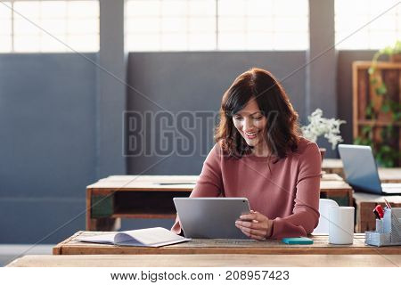 Attractive young businesswoman smiling while sitting alone at her desk in a modern office working online with a digital tablet