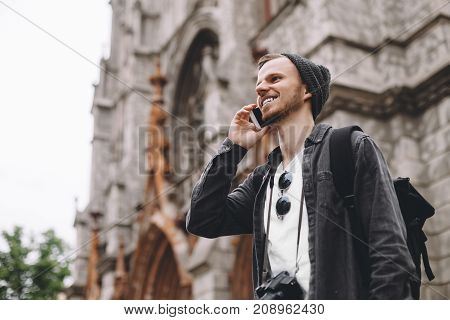 A picture of a young tourist standing near the old gothic church and talking on the phone. He is looking straight forward and smiling. The guy has almost reached the destonation place.