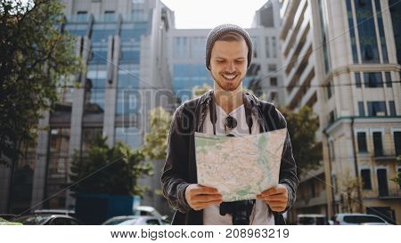 Beautiful and sunny picture of a handsome awesome guy standing outside on the street and looking to the map. There is a big building behind him. Young man has found a place to go so he is happy about that. Close up