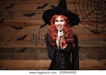 Halloween witch concept - Happy Halloween red hair Witch doing silence gesture with finger on her lips over old wooden studio background.