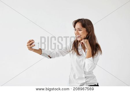 Charming happy young woman employee wearing formal white blouse taking selfie on smart phone smiling broadly at camera showing her perfect teeth having small break while working at office