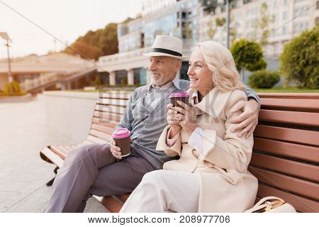 Two pensioners are sitting on a bench with a glass of coffee in their hands. They sit embracing and smiling. Pensioners are looking into the distance