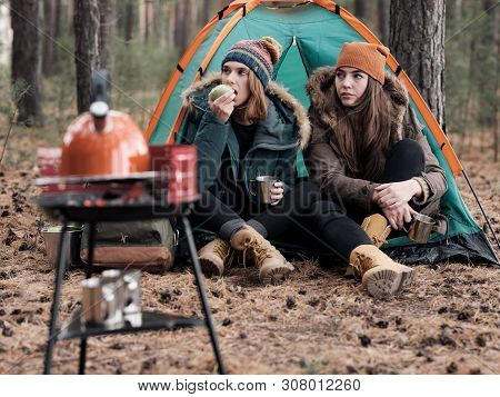 Girlfriends In Warm Clothes Sitting On Threshold Of Tent In Pine Forest, Eating Apple And Drinking T