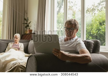 Front view of active senior Caucasian man sitting on sofa and using laptop while his wife using tablet in sofa in living room at comfortable home
