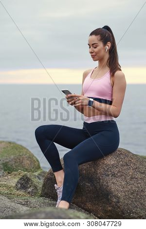 Happy Smiling Girl Is Chatting By Mobile Phone While Sitting On The Stone At Calm Seaside.