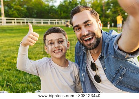Image of a happy young man having fun with his little brother or son outoors in park beautiful green grass take a selfie by camera showing thumbs up gesture.