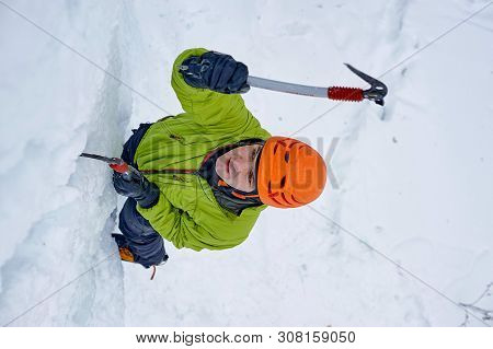 Alpinist Man With Ice Tools Axe In Orange Helmet Climbing A Lar