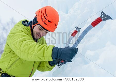 Athletic Alpinist Man In Orange Helmet And Ice Tools Axe Climbing A Large Wall Of Ice. Outdoor Sport