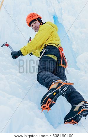 Athletic Alpinist Man In Orange Helmet And Ice Tools Axe Climbing A Large Wall Of Ice. Outdoor Sport