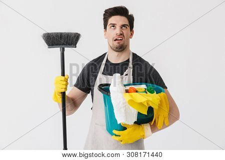 Handsome brunette houseman wearing apron standing isolated over white background, holding bucket full of detergents