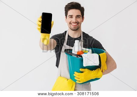 Handsome brunette houseman wearing apron standing isolated over white background, holding bucket full of detergents, showing blank screen mobile phone