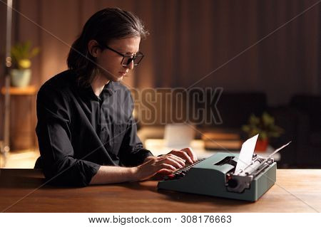 Confident Writer Working On A Typewriter, Seated At A Table, Over Blurred Interior. Copy Space.