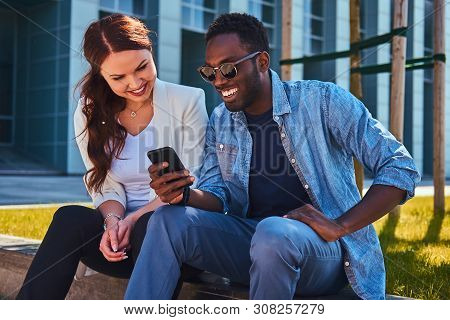 Happy Smiling Multicultural Couple Are Wathing Something On Mobile Phone While Sitting On The Grass 