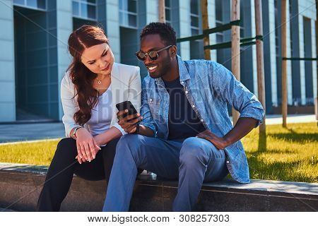 Happy Smiling Multicultural Couple Are Wathing Something On Mobile Phone While Sitting On The Grass 