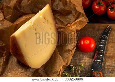 Piece Of Hard Cheese On Craft Paper, Thyme, Cheese Knife And Cherry Tomatoes On A Wooden Table