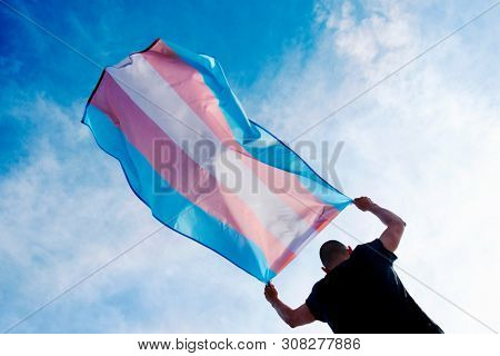 a young caucasian person, seen from behind, holding a transgender pride flag over his or her head against the blue sky