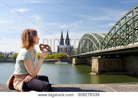 Young Woman Eats Traditional Pretzel Sitting On Embankment Of Rhine On Background Of Cologne Cathedr