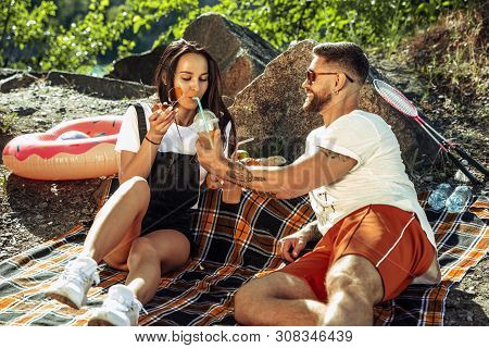 Young Couple Having Picnic At Riverside In Sunny Day. Woman And Man Spending Time On The Nature Toge