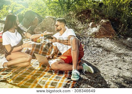 Young Couple Having Picnic At Riverside In Sunny Day. Woman And Man Spending Time On The Nature Toge