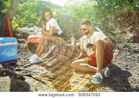Young Couple Having Picnic At Riverside In Sunny Day. Woman And Man Spending Time On The Nature Toge
