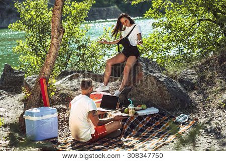 Young Couple Having Picnic At Riverside In Sunny Day. Woman And Man Spending Time On The Nature Toge